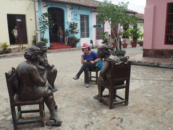 A woman in a pink hat sits among three statues of people also sitting down in a street square with yellow, pink and blue buildings behind them.