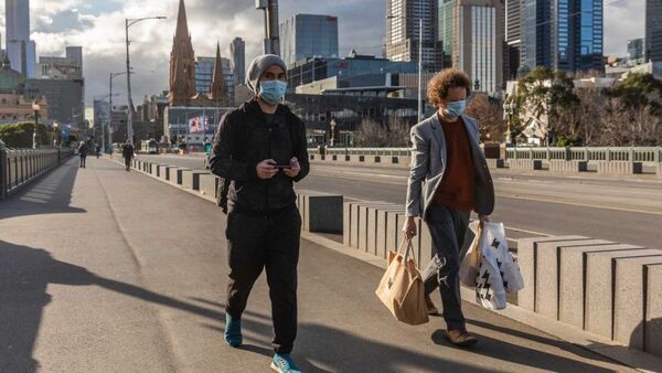 Two men wearing masks walk down a city street, one holds a phone, the other is carrying shopping bags