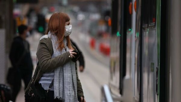 Young woman with long red hair wears a mask, scarf and coat while look at a tram
