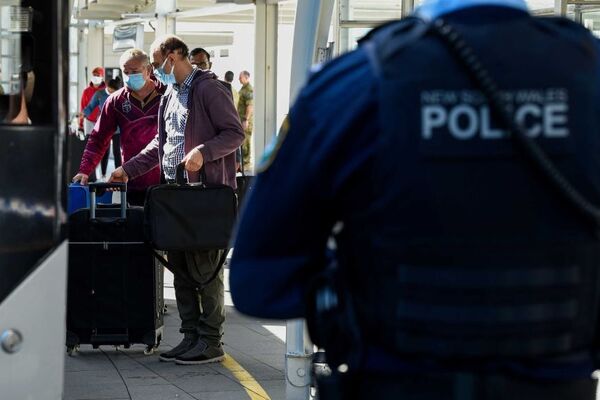 Back of police officer in foreground, two male passengers wearing masks, holding suitcases got through security in background
