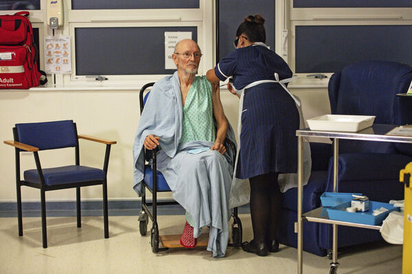 A man sits in a wheelchair as a woman stands over him and administers a vaccine in a hospital setting.