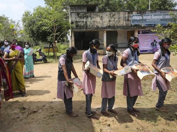 A line of girls hold boxes of packaged food and sweets. They wear uniforms and stand in front of a building, with older women to the left hand side.