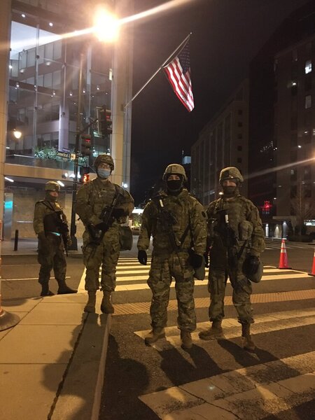 Four people are seen in army gear with guns strapped to their chests. An American flag is above them, and it is dark out.