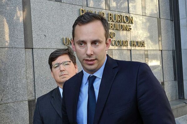 A white man stands in front of a court house building wearing a suit. Another man stands behind him.