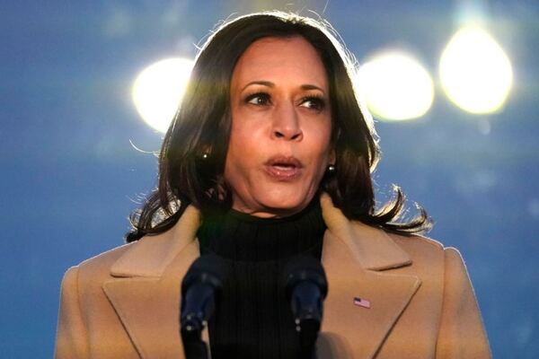 Vice President-elect Kamala Harris speaks during a COVID-19 memorial. She wears a camel coat with a US flag pinned to the lapel. Her hair is down, the sky behind her is mostly dark but two lights flare behind her head.