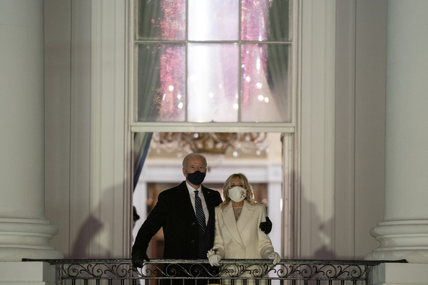 Joe Biden, with his arm around wife Jill Biden, stand on a balcony as they both look up to the sky. A reflection of fireworks is visible in the window above them