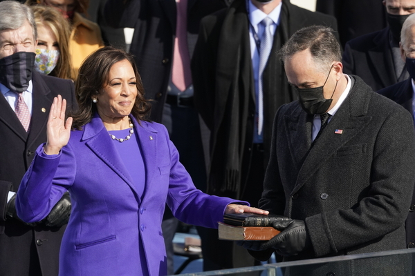 Kamala Harris, resplendent in purple, smiles proudly as she holds one hand up and the other on a stack of two bibles