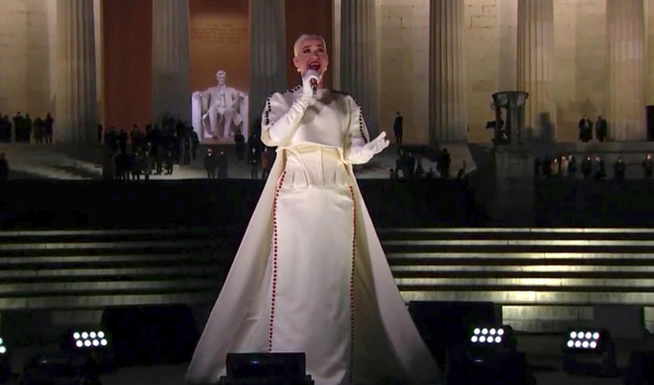 Katy Perry, in a white gown and gloves, sings into a microphone while standing on the steps of the Lincoln Memorial