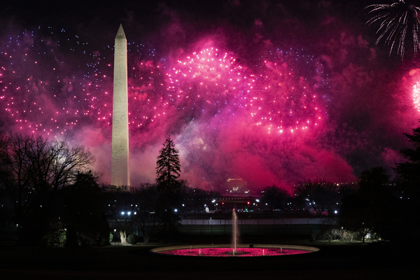 Pink fireworks explode across the night sky. The Washington Monument is visible, and the outlines of trees and city lights