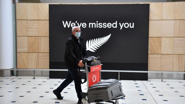A man in a mask pushes a suitcase trolley at Sydney airport, in front of a sign with silver fern that says: 