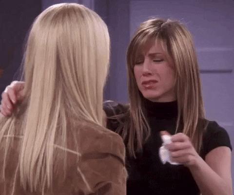 Rachel from Friends stands in front of a purple door and wall and holds a tissue in her left hand as she pulls Phoebe into an embrace and shuts her eyes, content.