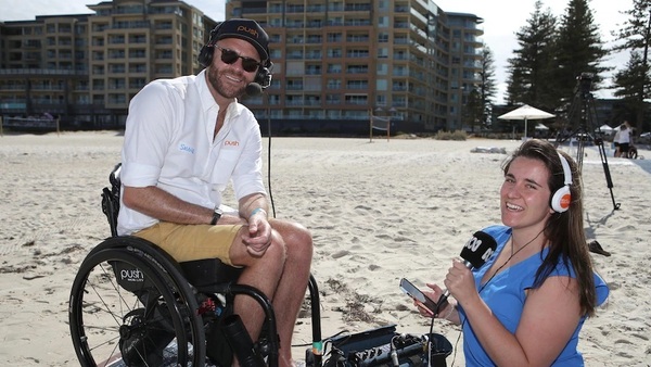 A man in a wheelchair and a kneeling woman conduct a radio interview on a beach.