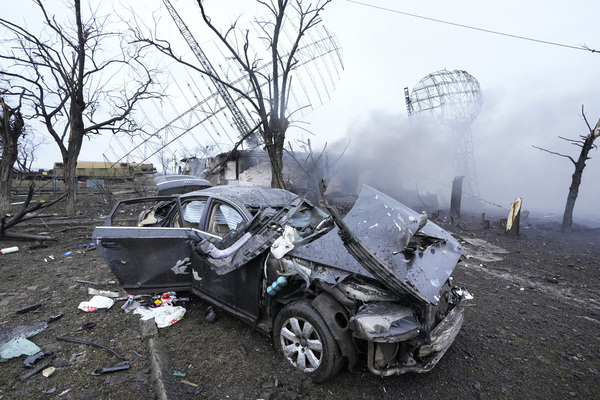 the wreckage of a car smokes in the foreground as twisted metal lies behind
