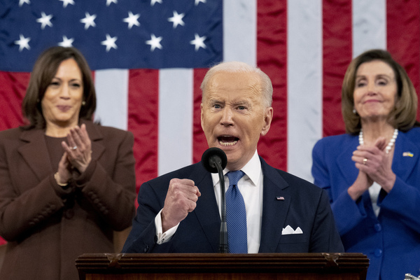 Joe Biden pumps his fist while standing at a microphone. Behind him Kamala Harris and Nancy Pelosi applaud in front of a US flag
