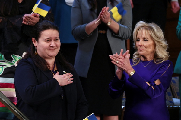 A woman stands with her hand clasped to her chest, while Jill Biden applauds beside her