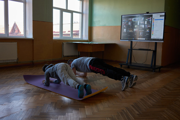 A young boy and his teacher in plank position in an empty classroom, a Zoom video call on the screen of a TV behind them