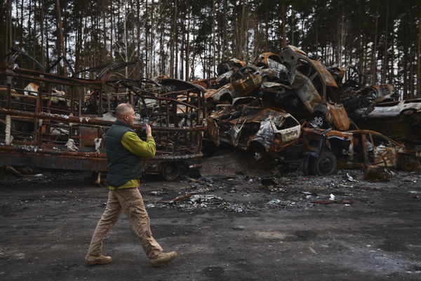 A man inspects the destroyed cars, piled high at the edge of a forest.