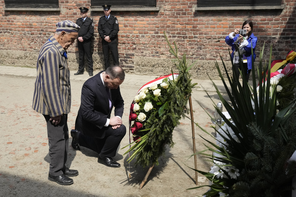 The polish president kneels, head down, in fromnt of a wreath. An elderly man in Auschwitz stripes stands next to him
