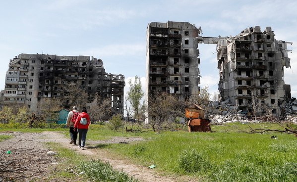 two people walk through a field, in the distance, two apartment blocks are partially collapsed
