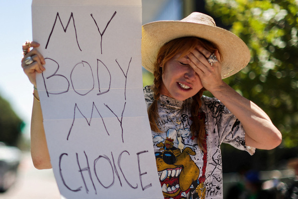A woman holds a MY BODY MY CHOICE sign while holding one hand up to her forehead in a look of despair